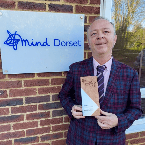Image shows John in a checked suit smiling and carrying an award. He's outside the Dorset Mind Office and our logo sign is behind him.