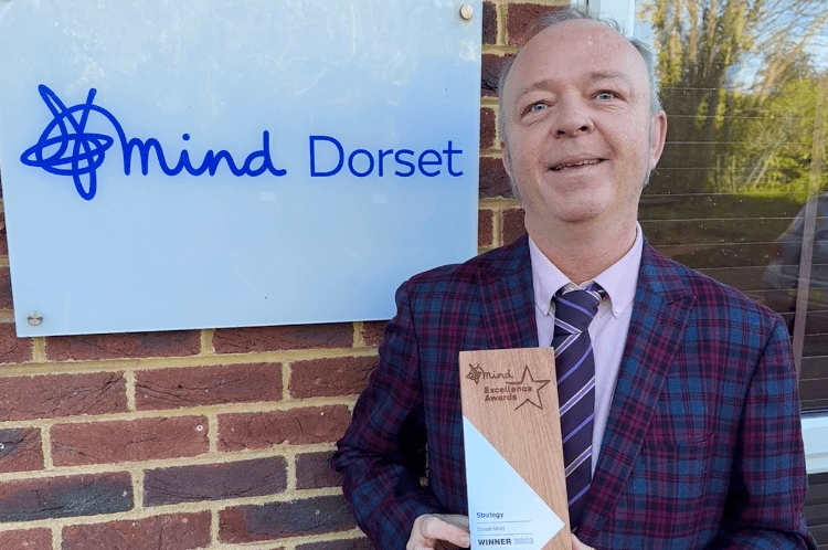 Mind Excellence Award Image shows John in a checked suit smiling and carrying an award. He's outside the Dorset Mind Office and our logo sign is behind him.
