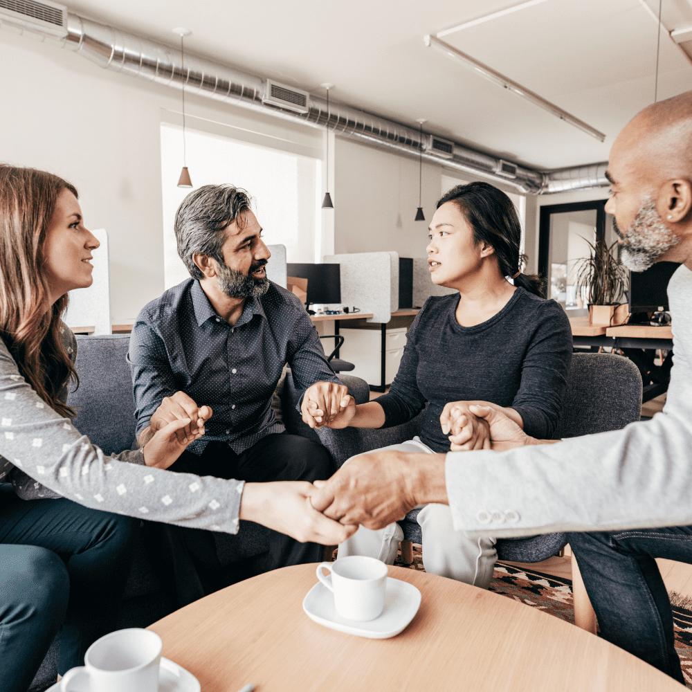 A group of four people around a coffee table, all holding hands and looking at on of them as they talk.