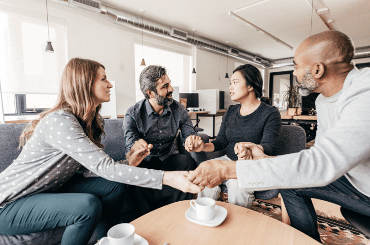 Celebrating World Mental Health Day Embracing Mental Well-Being A group of four people around a coffee table, all holding hands and looking at on of them as they talk.