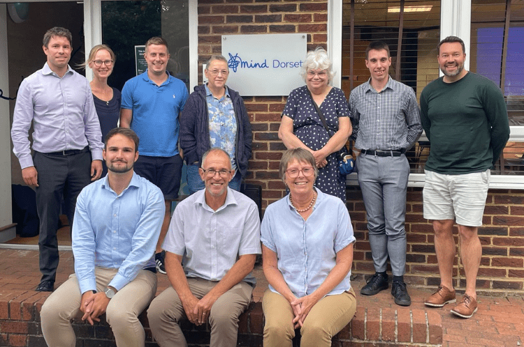 A group of people of various ages outside a building. 7 are standing and 3 are seated, all smiling for the camera. There is a plaque in the top row that carries Dorset Mind's logo.