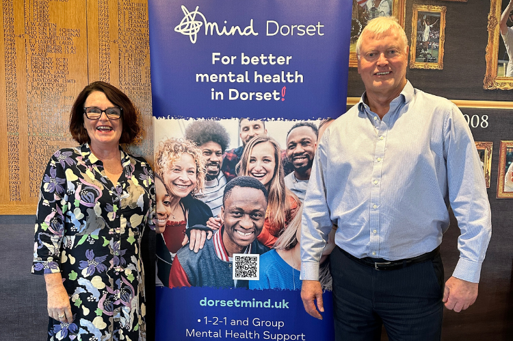 Dorset Mind CEO, Linda O'Sullivan smiles to the camera alongside The West Hants Club CEO, Peter Elviss. There's a Dorset Mind roller banner between them and they're standing in front of a wooden 'honours' board and photos of previoius Tennis Players.
