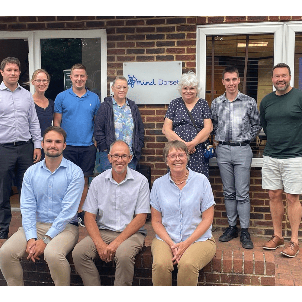 A group of people of various ages outside a building. 7 are standing and 3 are seated, all smiling for the camera. There is a plaque in the top row that carries Dorset Mind's logo.