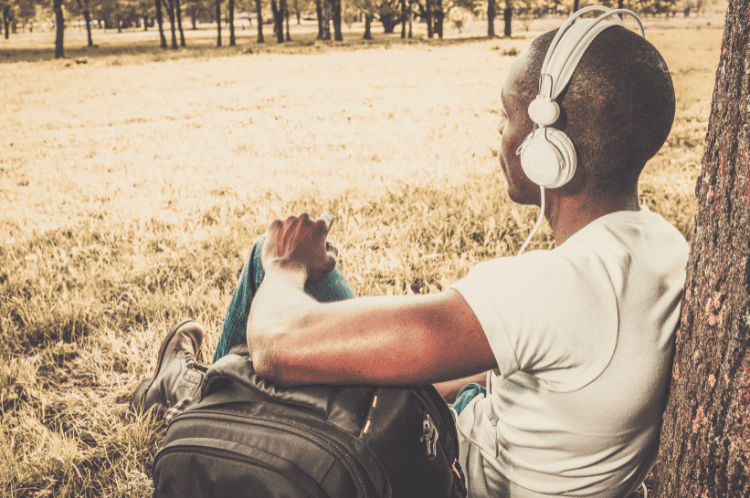 Untitled design (19) Black guy sat down against a tree putting on headphones, listening to music