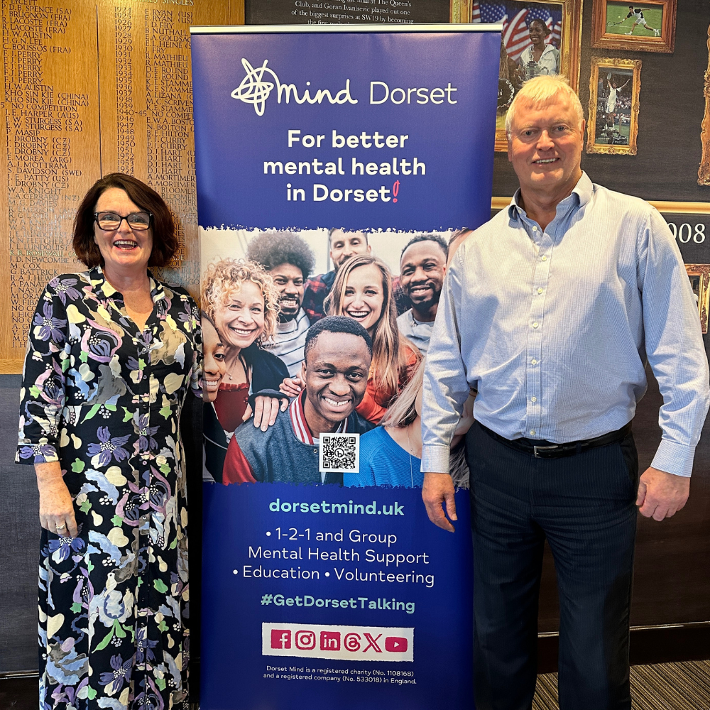 Dorset Mind CEO, Linda O'Sullivan smiles to the camera alongside The West Hants Club CEO, Peter Elviss. There's a Dorset Mind roller banner between them and they're standing in front of a wooden 'honours' board and photos of previous Tennis Players.