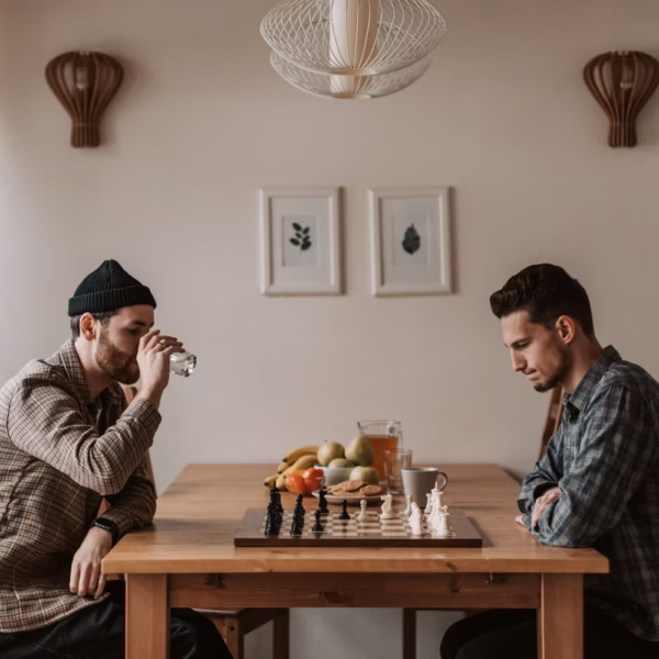 Two men sitting down at a table facing each other over a game of chess and drinks