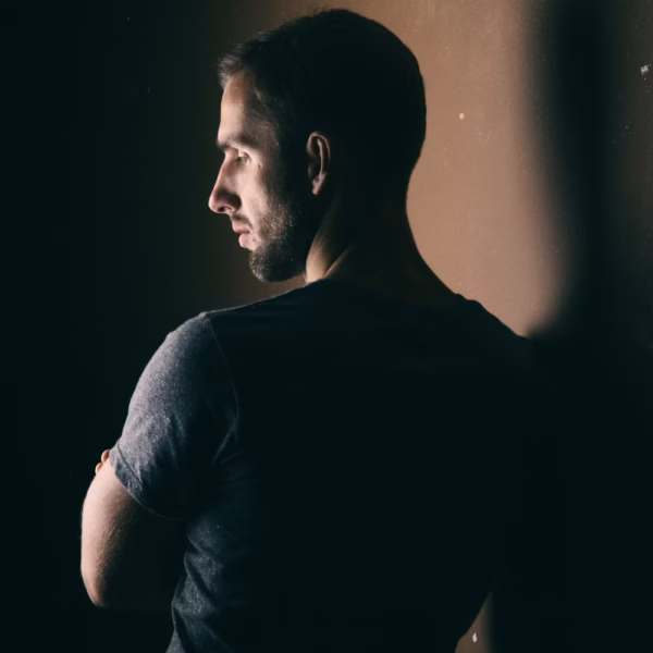 A youngish man standing in the shadows with a reflection of light on one side of his face.
