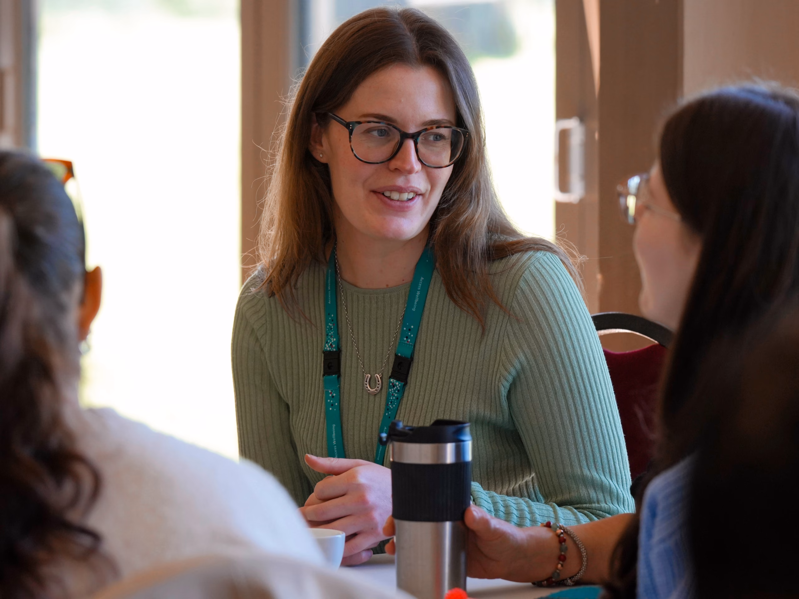 Female wellbeing coordinator smiling while running a drop-in session. She is wearing glasses and dressed in green.