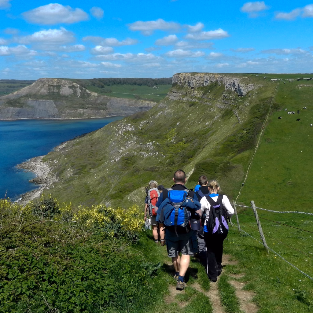 A group of people are walking the Jurassic Coast path, surrounded by green hills, with cliffs to the sea at their left.